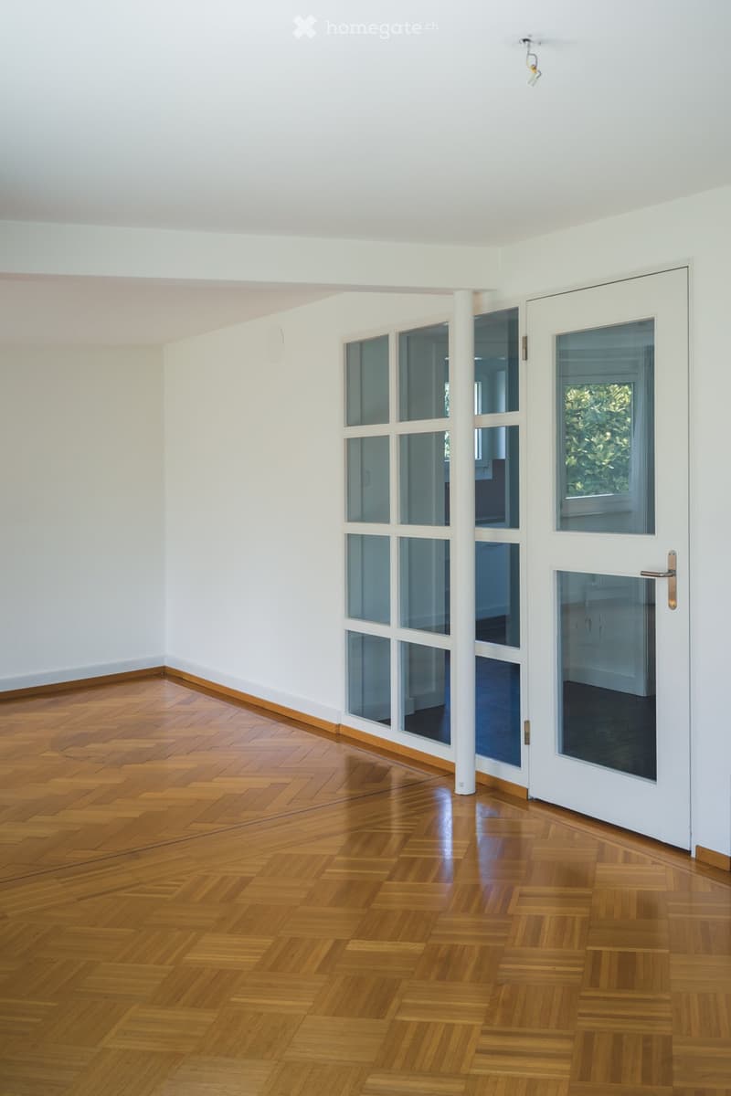 Empty room with wooden herringbone flooring, white walls, and large windows with glass panels. A white column and a white door are visible.