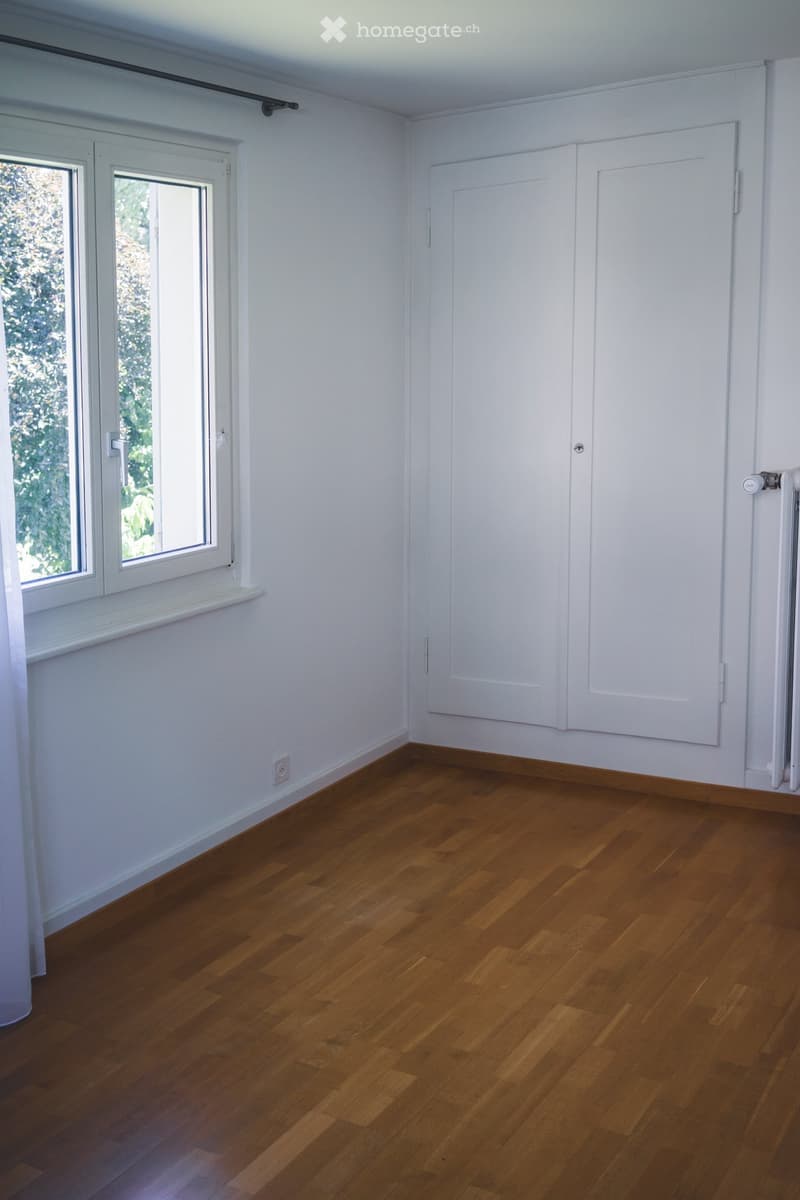 Empty room with wooden flooring, white walls, and large windows with white frames. There is a radiator on the right wall and a door with white panels.
