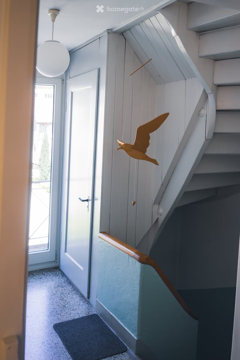 Staircase with wooden handrail, white walls, and a hanging bird decoration. A door and a window are visible in the background.