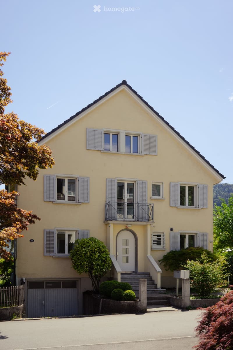 Two-story yellow house with white shutters, a balcony, and a garage. The house has multiple windows and a front door with steps leading up to it.