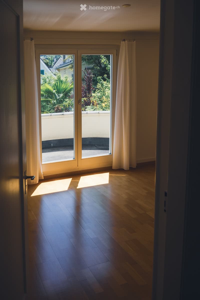 Empty room with wooden flooring, large sliding glass doors leading to a balcony, and white curtains on either side.