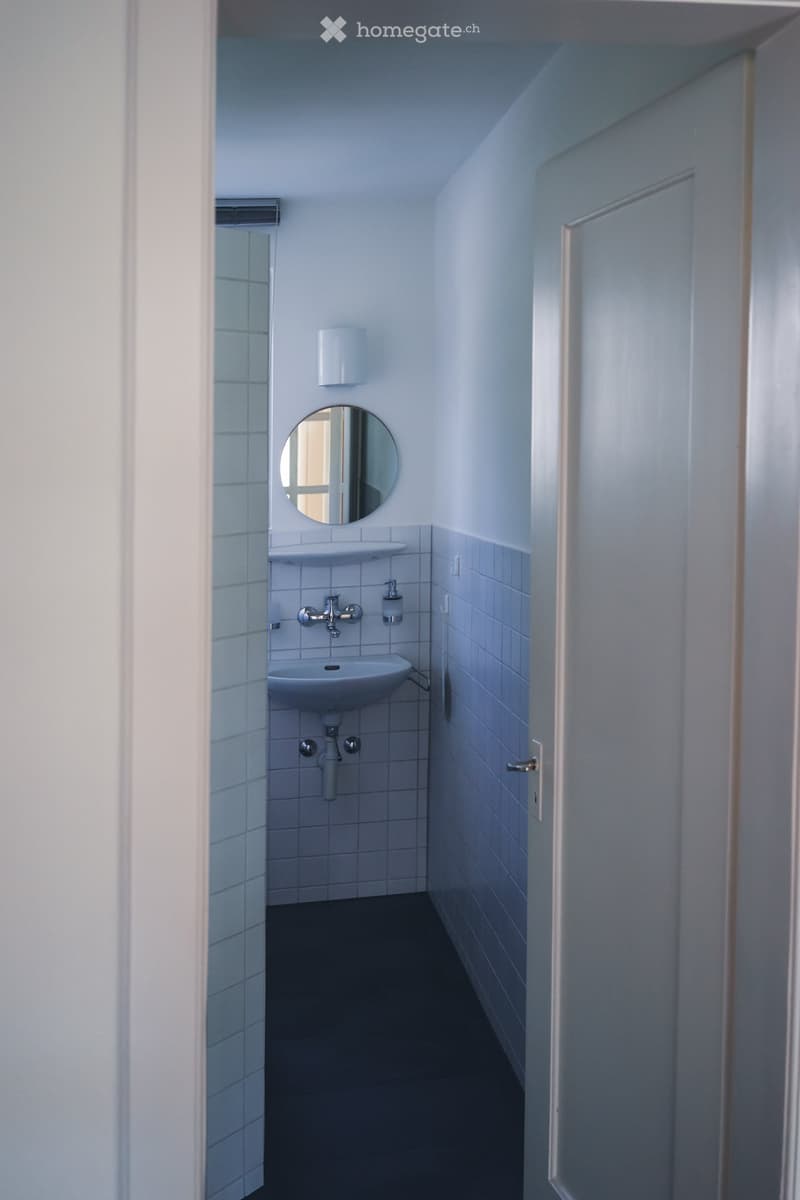 Small bathroom with white tiled walls, a white sink, a round mirror, and a soap dispenser. The floor is dark, and the space is well-lit.