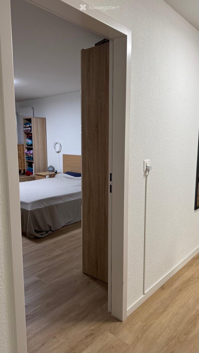 A modern hallway with light wooden flooring and white walls, leading to a bedroom with a bed, nightstand, and wardrobe.