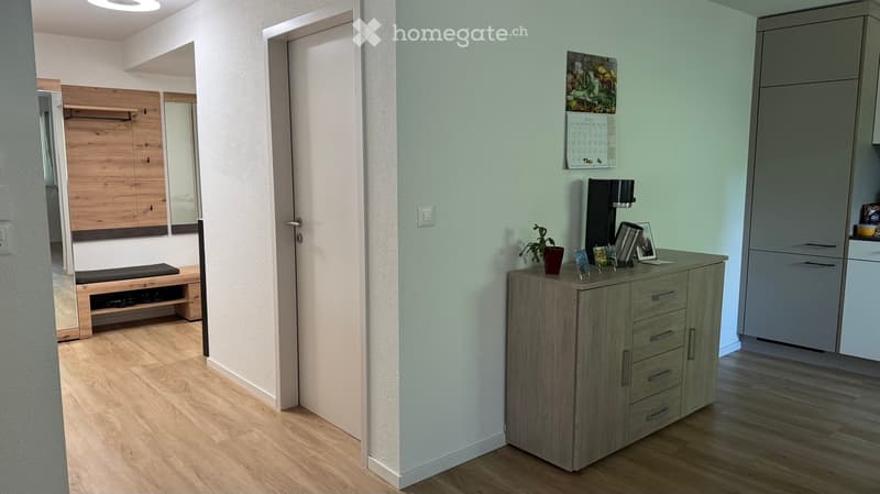 A modern hallway with light wooden flooring, a grey cabinet with drawers, a small bench, a mirror, and a calendar on the wall.