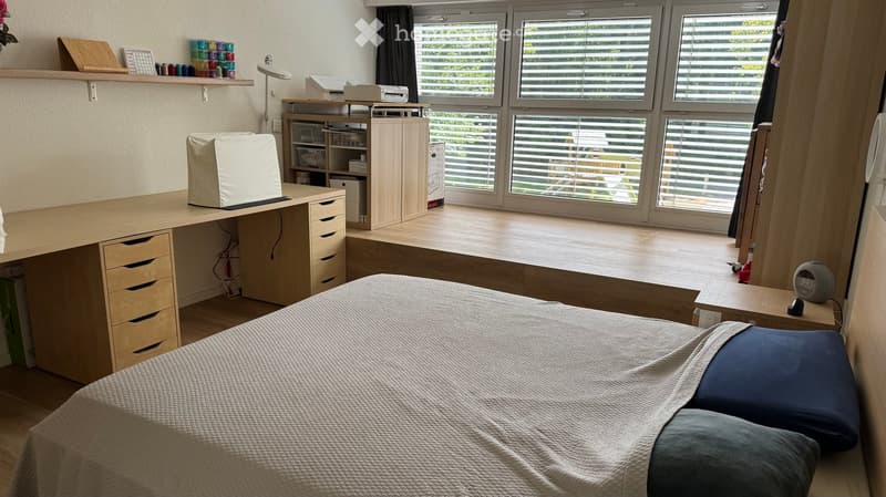 Well-lit bedroom with a bed, desk, nightstand, and large windows with blinds. The room features a modern, minimalist design with light wood furniture and a cozy bed.