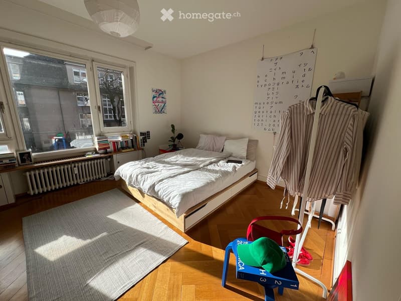 Cozy bedroom with a wooden bed, white bedding, a small table with books and a plant, a chair with a green cap, and a clothing rack with hanging garments.