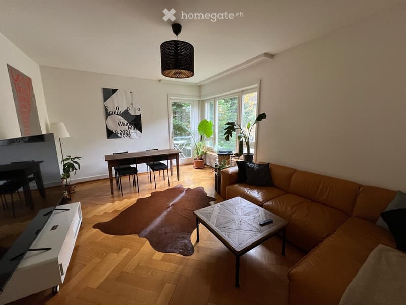 Modern living room with a leather sofa, coffee table, dining area, large windows, and decorative elements including a cowhide rug and artwork.