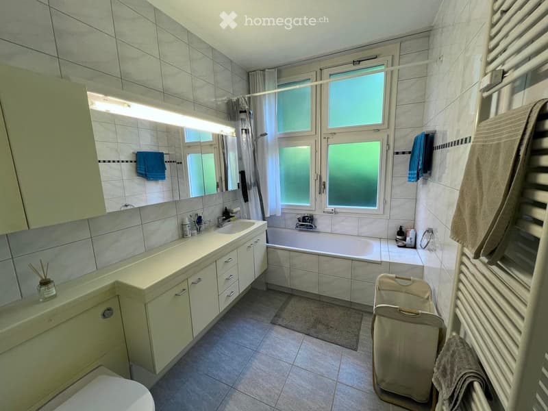 Bathroom with a sink, bathtub, mirror, radiator, and towels. The room features tiled walls and floor, a window with frosted glass, and a cabinet with drawers.