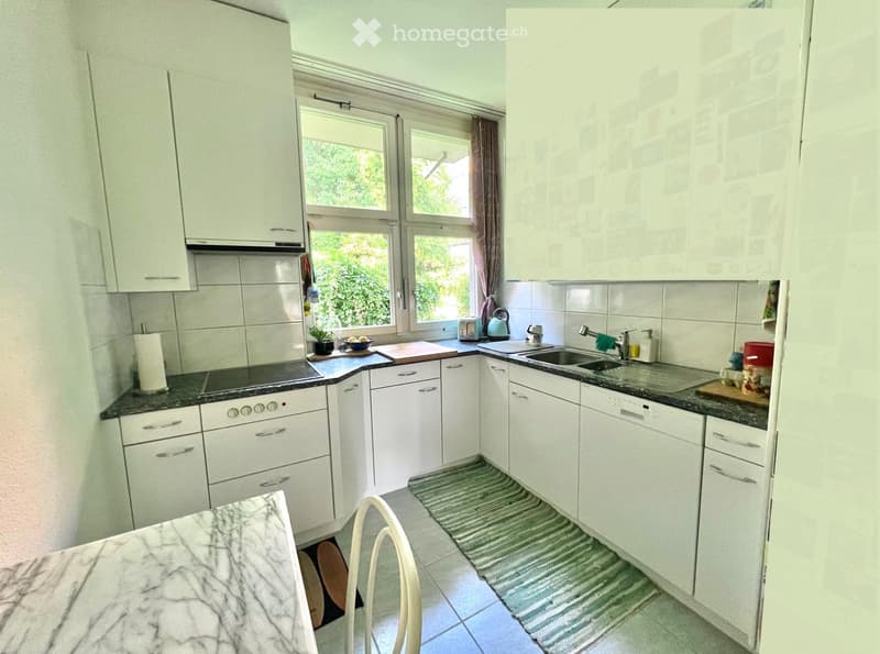 Bright kitchen with white cabinets, granite countertops, and stainless steel appliances. Features a sink, oven, toaster, teapot, and a small dining area with a marble table and chair.