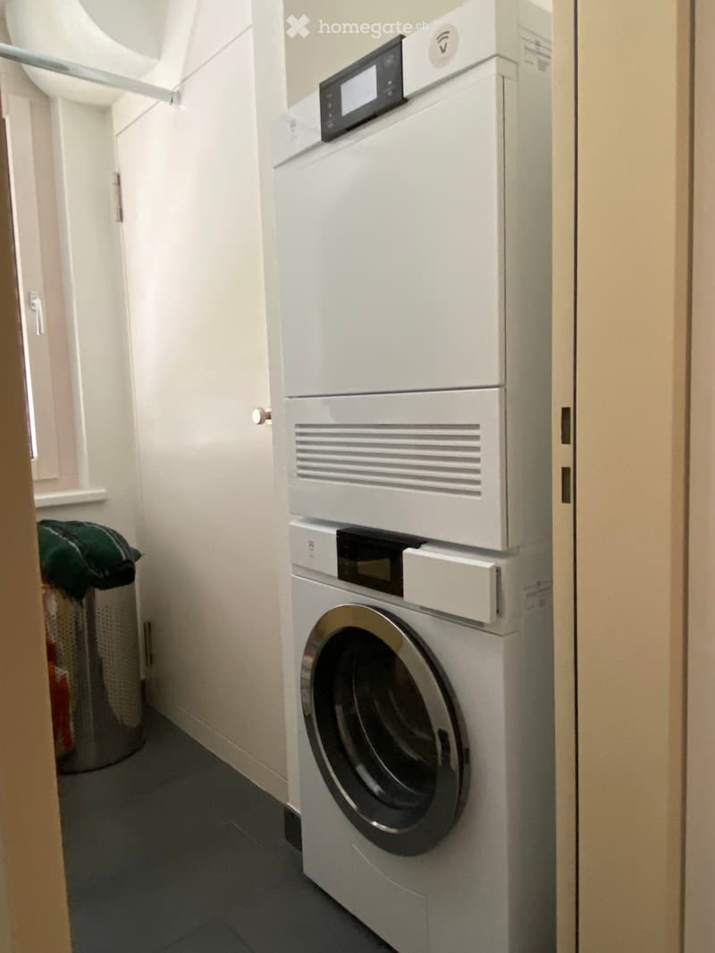 A compact bathroom with a stacked washing machine and dryer unit, a trash bin, and light-colored walls.