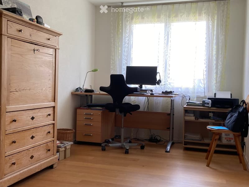 A well-lit study or home office with a wooden floor, a large window with sheer curtains, a desk with a computer, a chair, a lamp, a printer, and a small wooden stool with a backpack on it. A wooden dresser with drawers is on the left side.