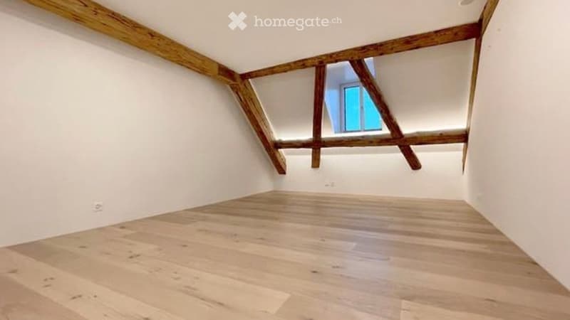 Empty attic room with wooden beams and a skylight window, featuring light wooden flooring and white walls.