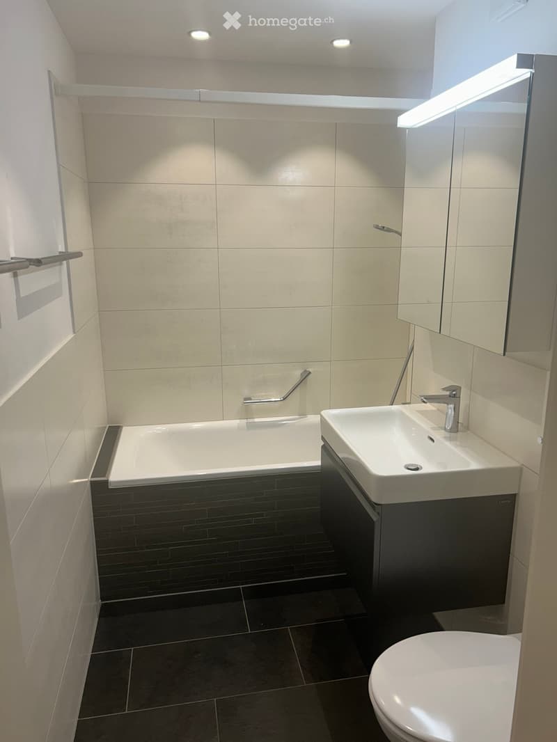 Modern bathroom with a bathtub, sink, toilet, and mirror. Features light-colored tiles and dark flooring.
