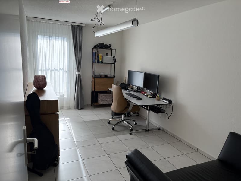 Modern office space with a desk, ergonomic chair, dual monitors, keyboard, mouse, shelf with books and decorative items, and a guitar leaning against the wall.