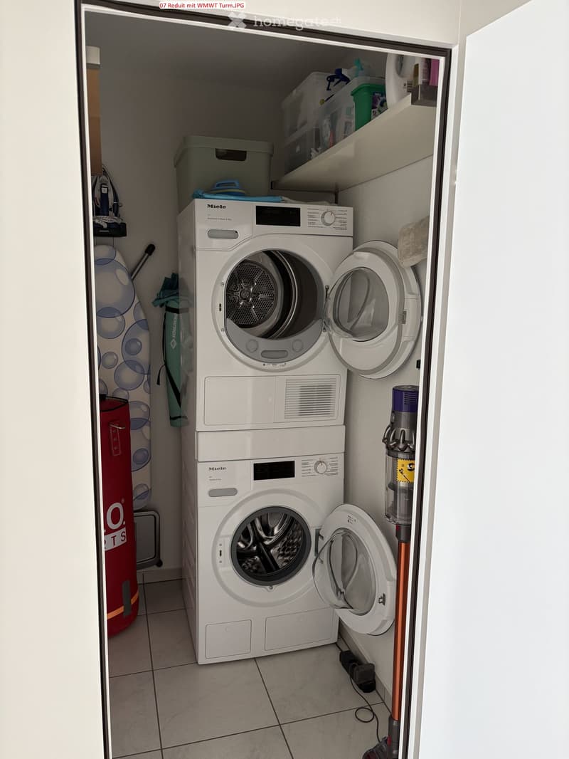 A compact laundry area with a stacked washing machine and dryer unit, shelves with storage boxes, and a vacuum cleaner stored nearby.