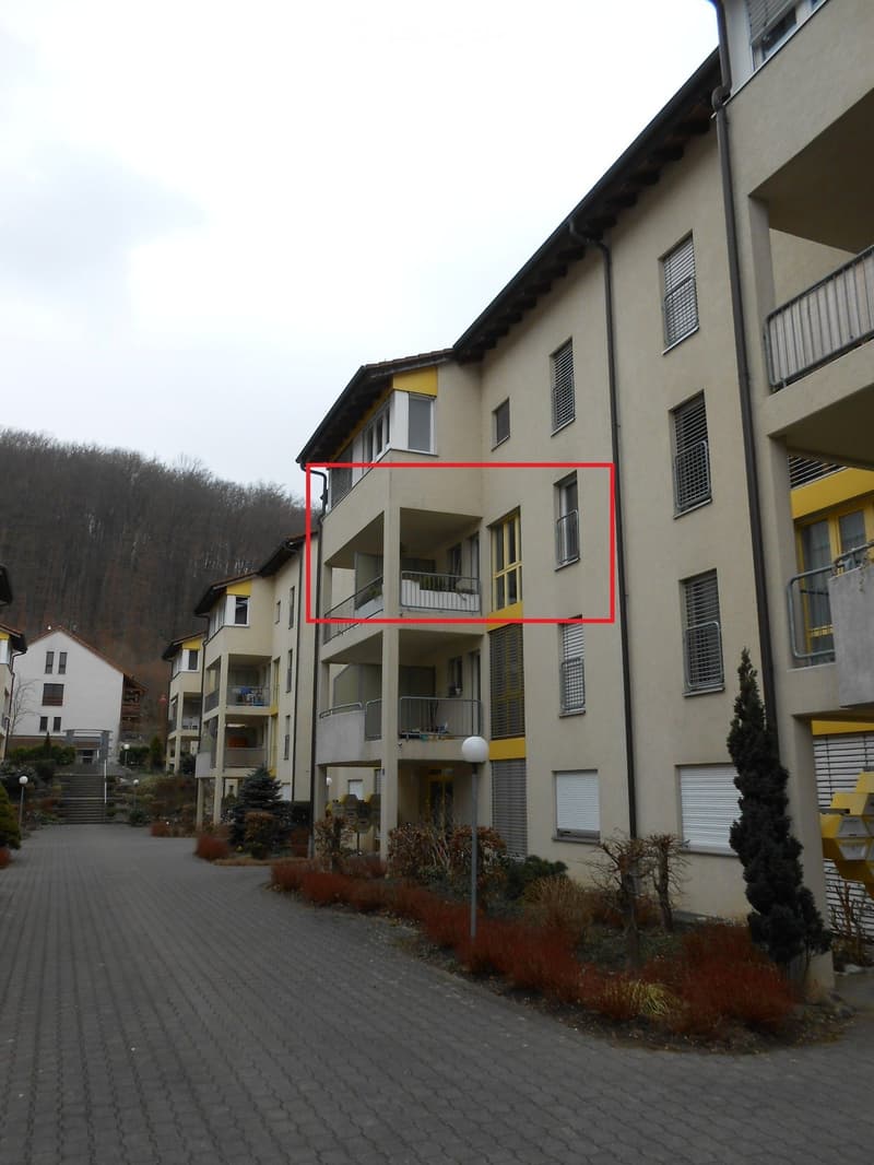 Multi-story residential building with balconies, yellow accents, and surrounding greenery.