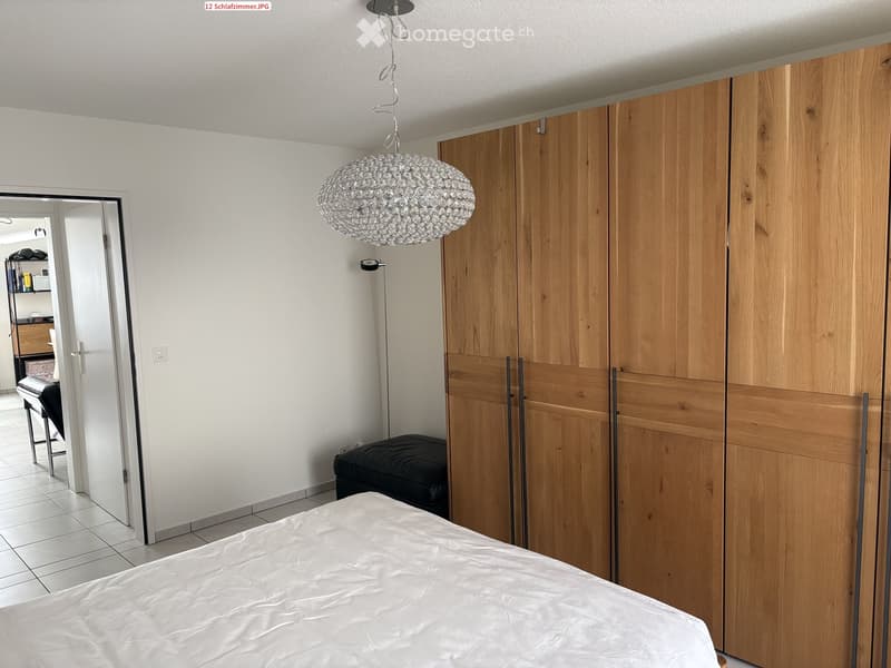 Modern bedroom with a large wooden wardrobe, a round crystal chandelier, a bed with white bedding, and a black leather stool.
