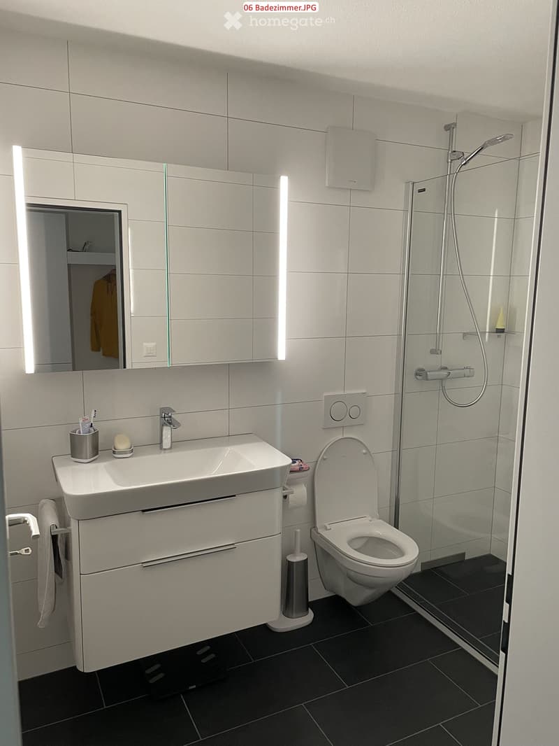 Modern bathroom with white tiles, a large sink with a modern faucet, a toilet, a glass-enclosed shower, and a large mirror with built-in lighting. The floor is covered with dark tiles.