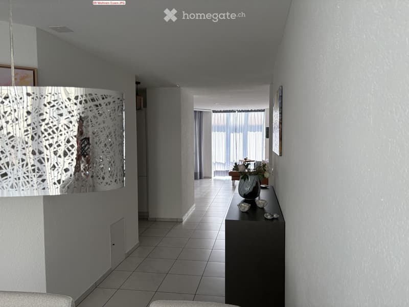 Modern hallway with tiled floor, white walls, and a decorative console table with a vase and small decorative items. A chandelier hangs from the ceiling, and a framed picture is visible on the wall.