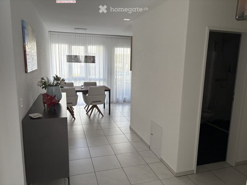 Modern dining area with a sleek black cabinet, a decorative vase with flowers, a small red sculpture, and a dining table with four chairs. The room features tiled flooring and large windows with sheer curtains.