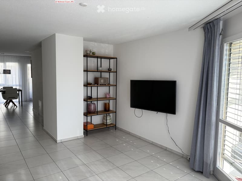Modern living room with tiled floor, large window with blinds, mounted TV, shelf with decorative items, and a small dining area with a table and chairs.