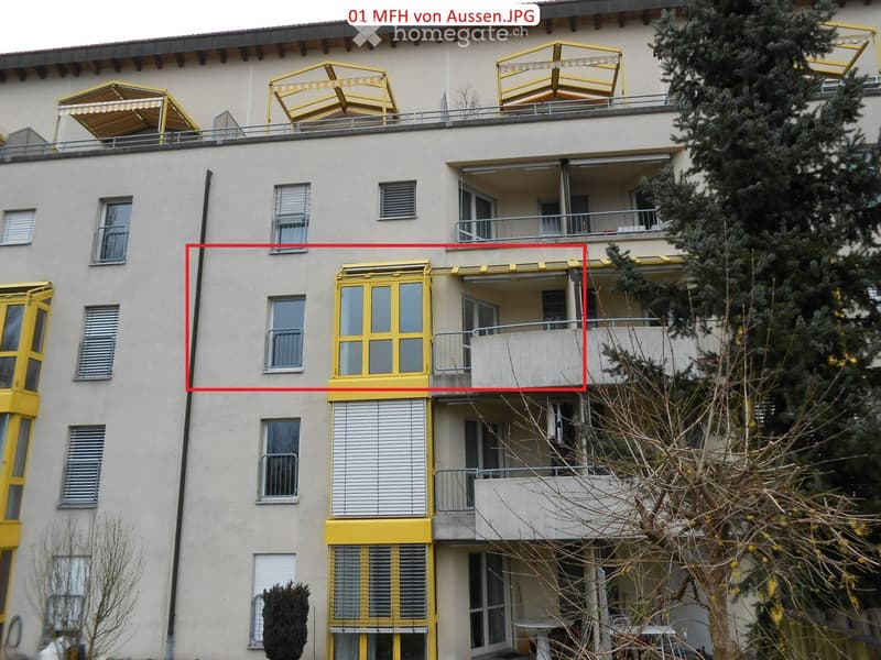 Multi-story residential building with balconies, yellow railings, and windows. The building has a light-colored facade and is surrounded by trees.