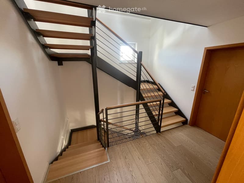 Modern staircase with wooden steps and metal railing, leading to an upper level. Light-colored walls and wooden flooring.
