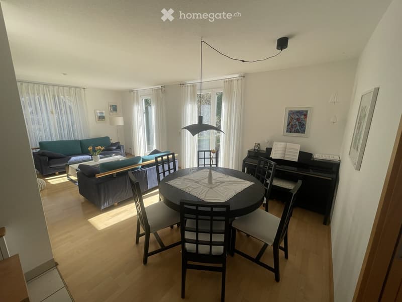 Bright living and dining area with wooden flooring, large windows with white curtains, a dark blue sofa, a round dining table with chairs, a piano, and wall art.
