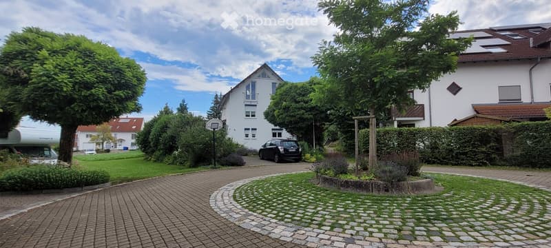 Residential area with multiple houses, well-maintained gardens, and a paved driveway leading to a central house.