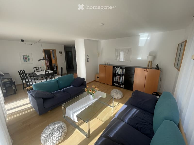 Modern living room with a blue sectional sofa, glass coffee table, two round stools, a wooden cabinet, dining area with chairs and table, and a bookshelf with books.