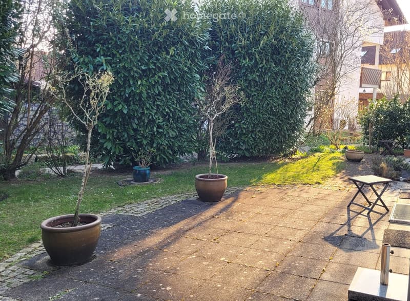 Garden area with paved stone pathway, potted plants, small table, and surrounding greenery.