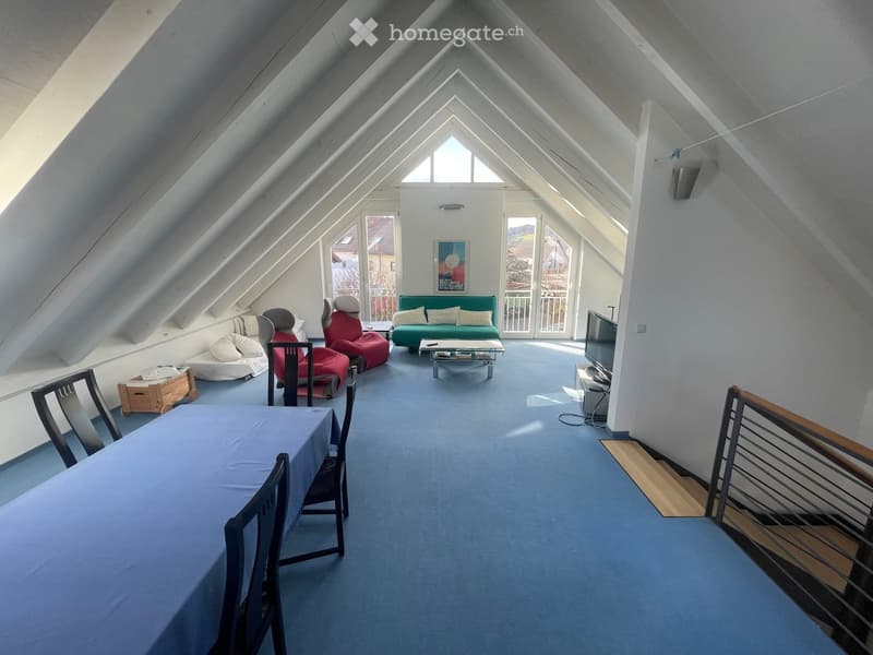Bright attic room with slanted ceilings, large windows, and skylights. Features a green sofa, red bean bag chairs, a dining table with chairs, and a TV on a stand.