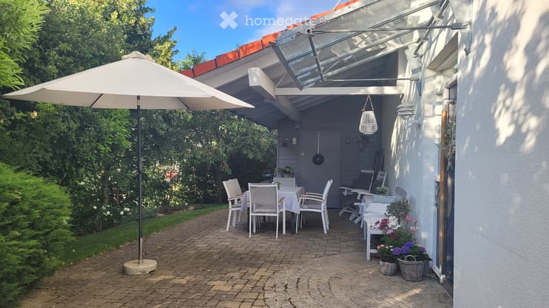 Outdoor patio area with a table, chairs, umbrella, and flower pots, surrounded by greenery and a covered entrance.