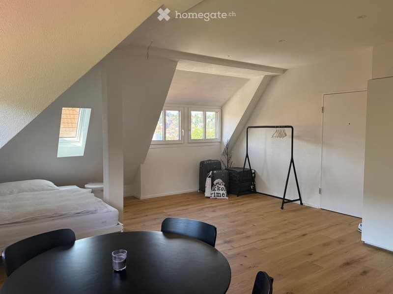 Bright attic bedroom with wooden flooring, slanted ceilings, and large windows. Features a bed, round table with chairs, and a coat rack.
