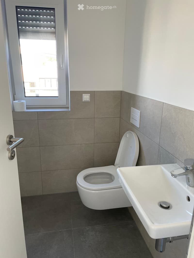 Modern bathroom with a toilet, sink, and window. The walls are tiled in a two-tone design with light upper tiles and darker lower tiles.