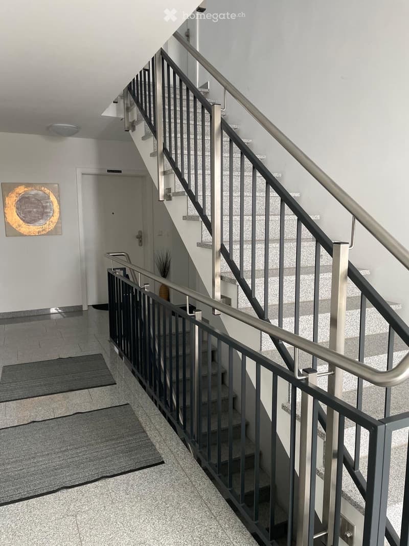 Modern hallway with a staircase featuring a metal railing, grey tiled floor, white walls, and a painting on the wall.