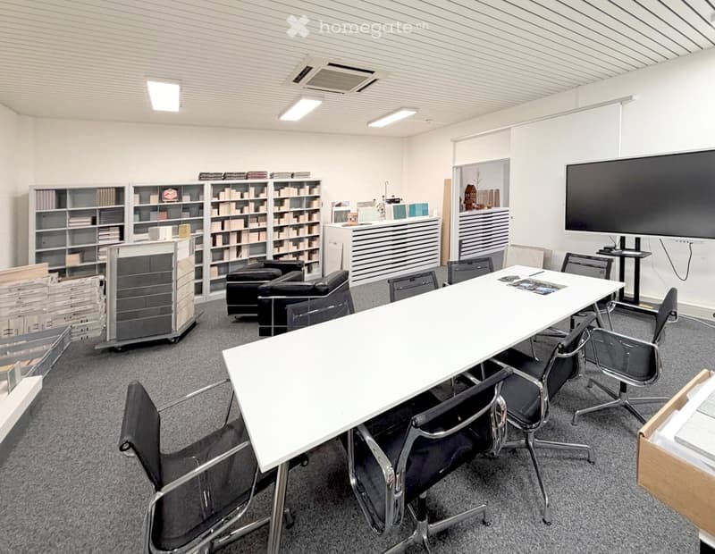 Modern office or meeting room with a large white table surrounded by black chairs, a screen on the wall, bookshelves, and a small sofa area.