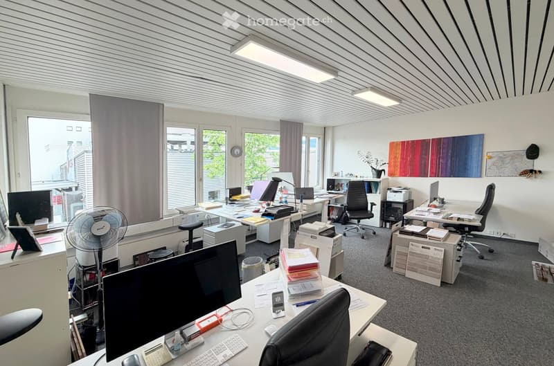 Office space with multiple desks, chairs, computer monitors, keyboards, printers, a fan, a clock, a plant, artwork, and a laptop.