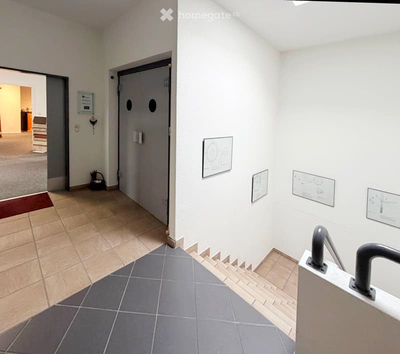 A clean, well-lit hallway with tiled floors, white walls, and a staircase leading upwards. There are several informational posters on the walls and a small basket near the entrance.