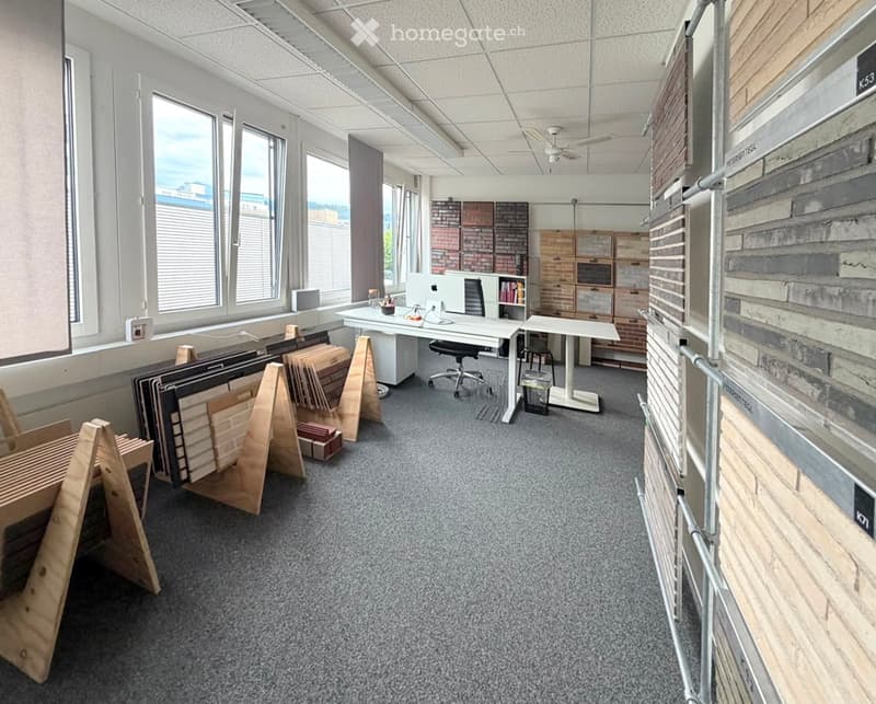 Office space with desks, chairs, a laptop, bookshelf, and storage units. Large windows provide natural light, and sample materials are displayed on the floor.