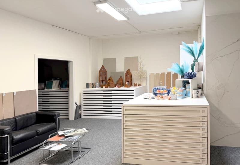 A modern, well-lit room with a sleek white desk, a black leather sofa, a glass coffee table, and decorative items including model houses and a floral arrangement.