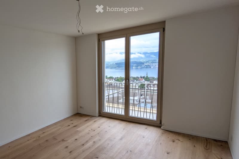 Empty room with wooden flooring, large sliding glass doors leading to a balcony with a scenic view of a lake and mountains.