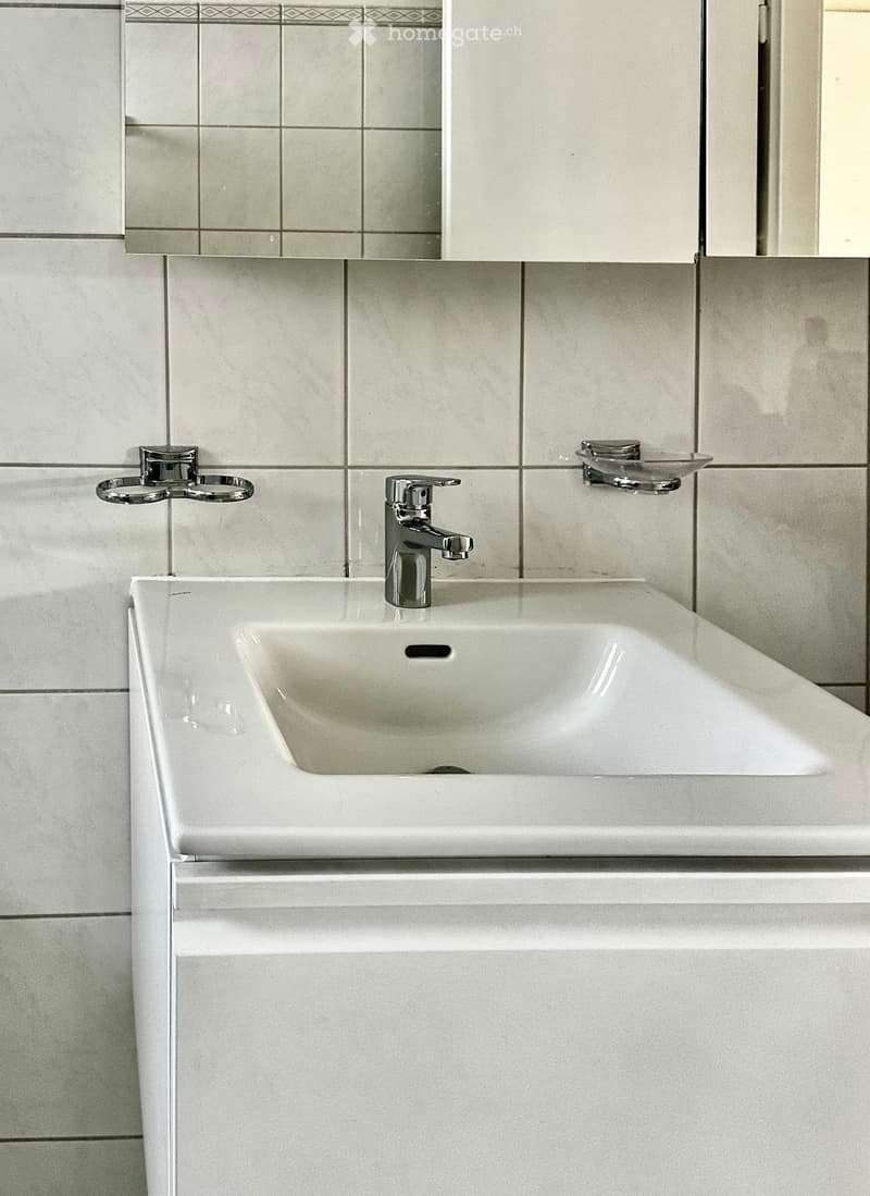 Modern bathroom with a white sink, chrome faucet, and a large mirror above it. The walls are tiled in light grey with decorative borders.