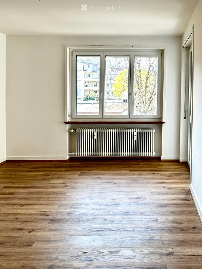 Empty room with wooden flooring, white walls, large window with radiator below, and a door on the right side.