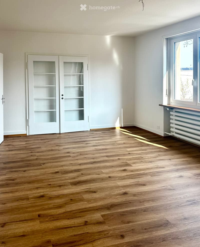 Empty room with wooden flooring, white walls, a large window, and a white door with glass panels.