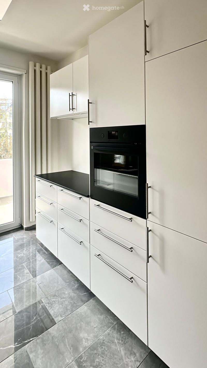Modern kitchen with white cabinets, black countertop, and built-in oven. Grey tiled floor and large window.