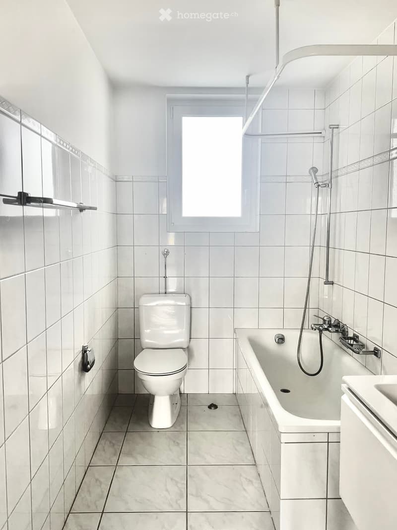 Modern bathroom with white tiles, a toilet, a bathtub with a showerhead, and a window providing natural light.