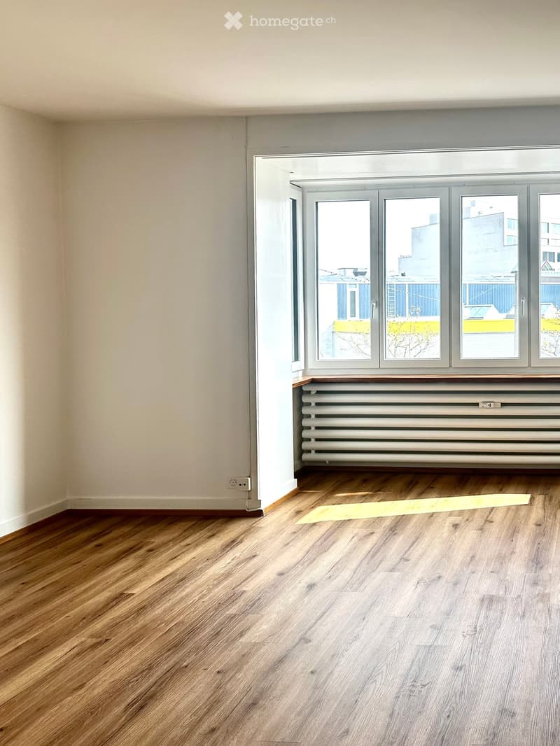 Empty room with wooden flooring, large window with radiator below, and a small yellow mat on the floor.