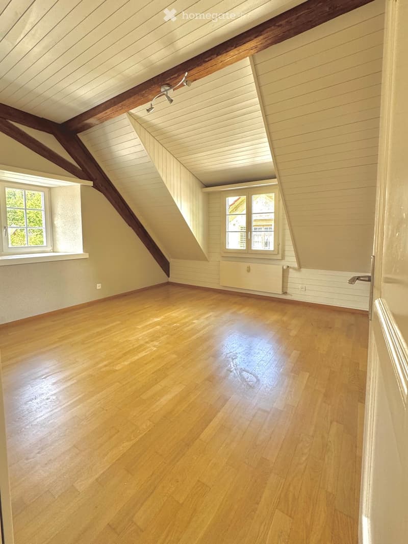Empty attic room with wooden floors, slanted ceilings, exposed wooden beams, and multiple windows letting in natural light.