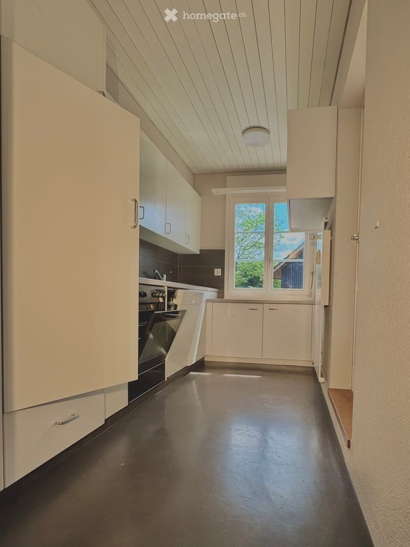 Modern kitchen with white cabinets, dark floor, and large windows overlooking greenery.